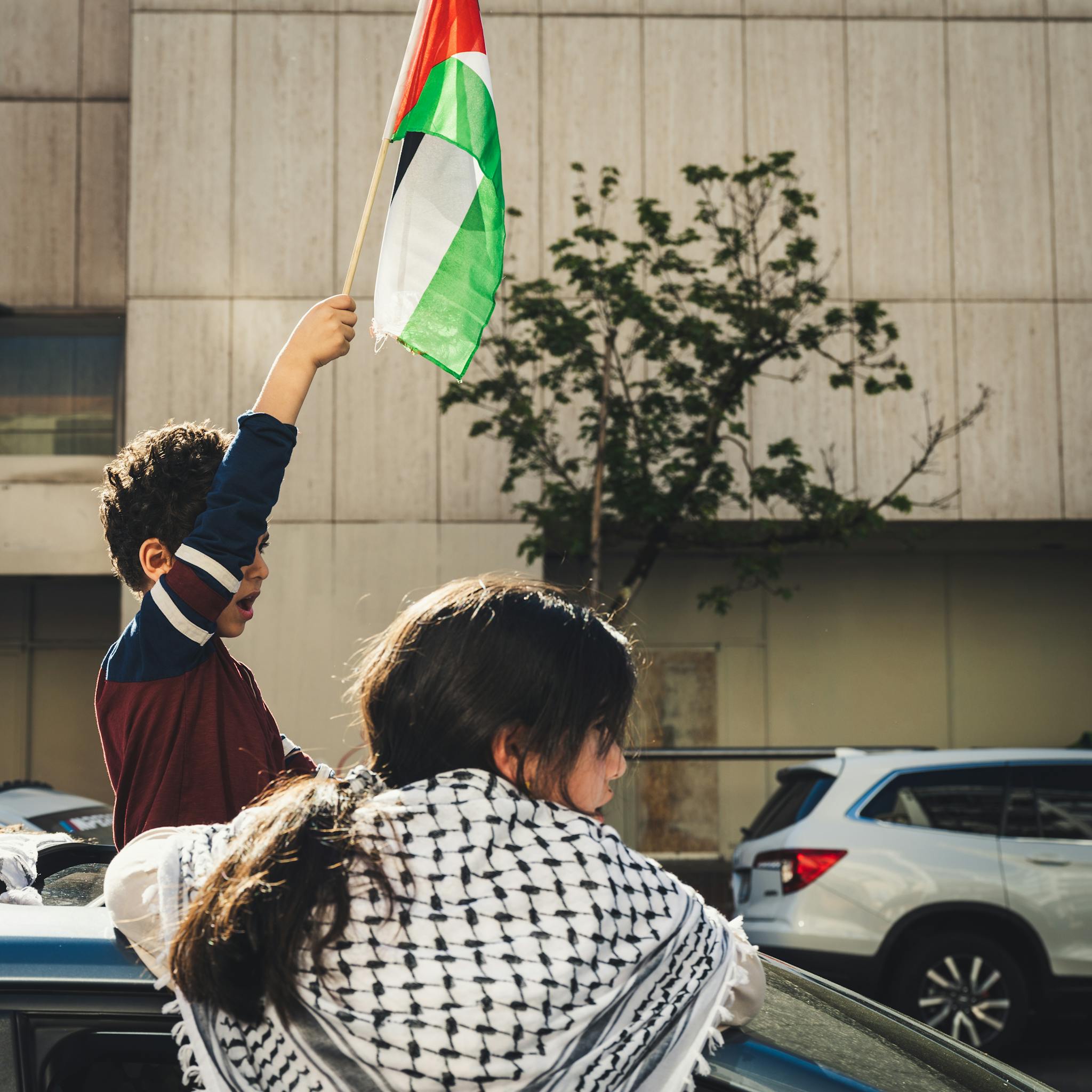 A young boy waves a Palestinian flag during an outdoor demonstration with cars.